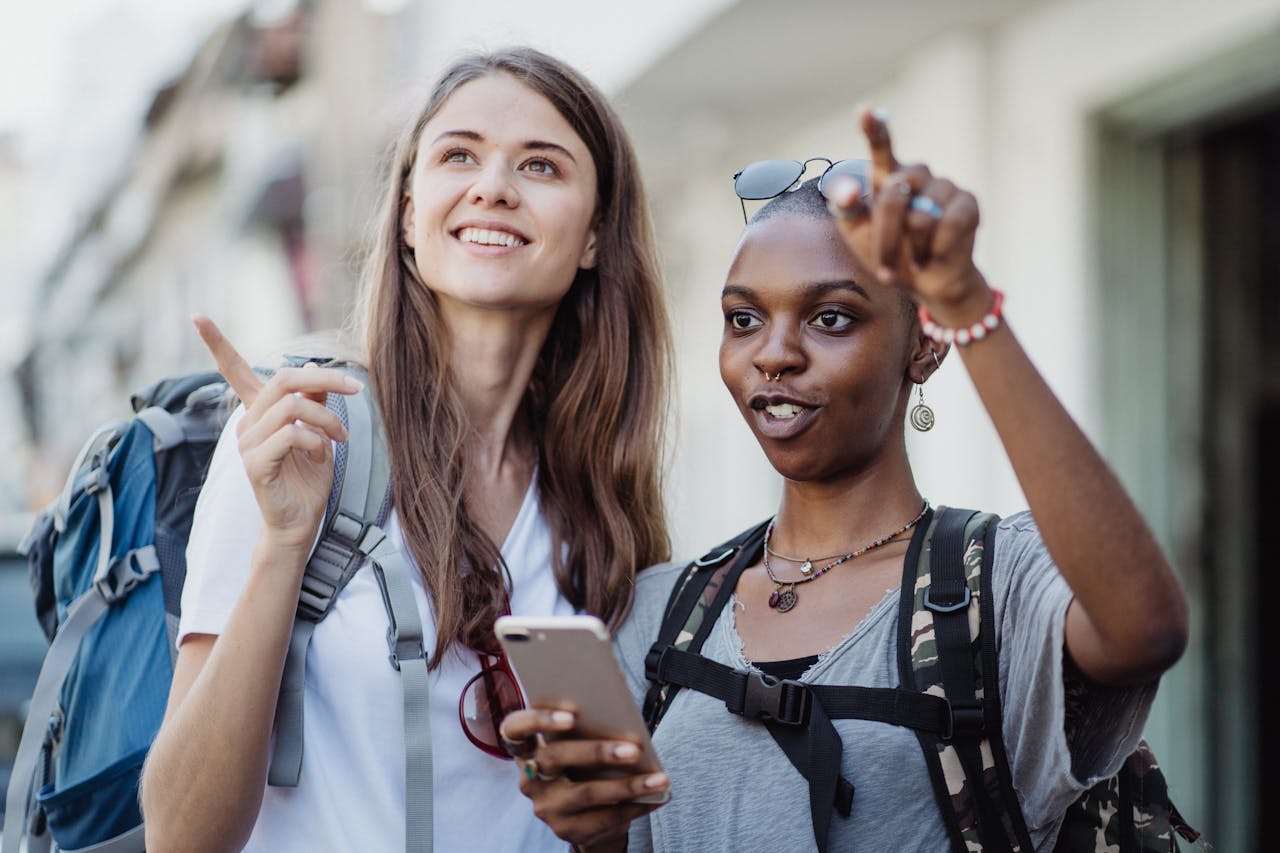 featured-images-01 Two smiling women with backpacks navigating a city using a smartphone, embracing adventure and travel.